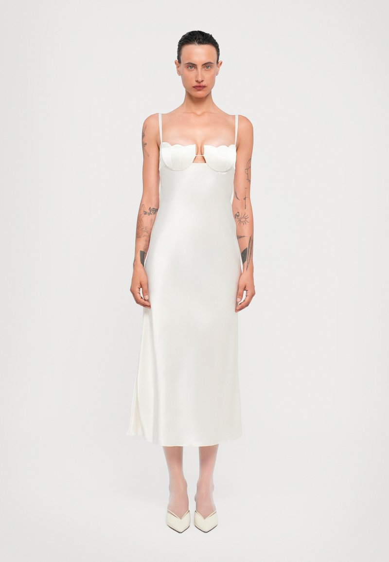 Witte zijden midi-jurk met gegolfd decolleté, dunne bandjes en een zijsplits. Model draagt crème puntige schoenen, met zichtbare lichaamstattoos.
