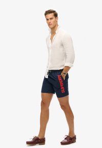 Camisa de lino blanca de manga larga, pantalones cortos de natación azul marino con estampado rojo de "SUPERDRY", zapatos de barco de cuero y un reloj con cara redonda.
