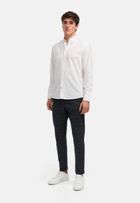 Chemise blanche à boutons et manches longues, associée à un pantalon écossais sombre et des baskets blanches. La chemise présente un col classique et des détails subtils.