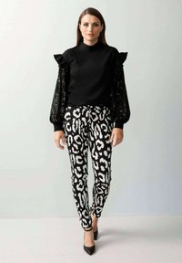 Schwarzes Oberteil mit Rüschenärmeln, kombiniert mit eng anliegenden schwarzen und weißen Leopardenmuster-Hosen. Das Model trägt schwarze High Heels.