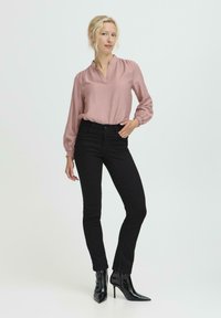 Rosenfarbene Bluse mit V-Ausschnitt und gerafften Ärmeln, kombiniert mit schwarzen Skinny Jeans und glänzenden schwarzen High Heels, vor einem hellen Hintergrund.