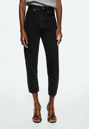 Person iført sorte, højtaljede cropped jeans med knap- og lynlåslukning, kombineret med sorte sandaler med hæl og stropper.