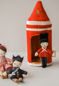 Červený látkový domeček na hraní s špičatou střechou a bílými pruhy. Tři textilní panenky: dívka v růžovém, voják v červené a chlapec v tmavě modrém.