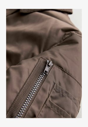 Hnědá izolační bunda s hladkou texturou, zapínáním na zip a nášivkovou kapsou na rukávu se šicími akcenty.
