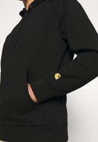 Svart hoodies med känguruficka, ribbad mudd och en guldlogotyp på ärmen. Mjukt material med en avslappnad design.