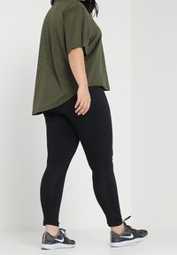 Oliivinvihreä verkkopaita lyhyillä hihoilla, yhdistettynä mustiin legginsseihin ja kuviollisiin urheilukenkiin. Pyöreä helma ja rento istuvuus.