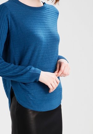 Woman wearing a blue ribbed sweater and black skirt, adjusting sweater hem with both hands against a neutral background.