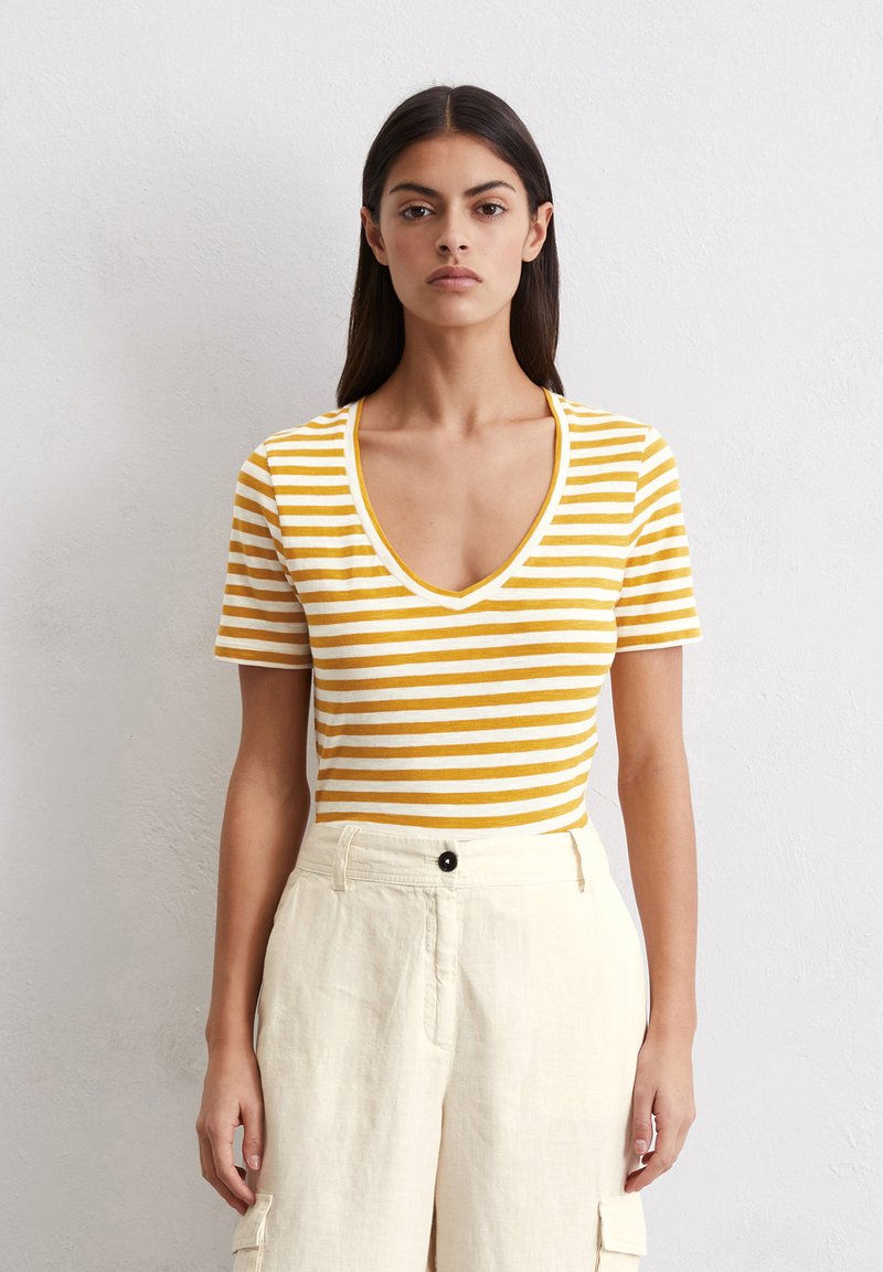 Gelb-weißer gestreifter Kurzarm-Oberteil mit V-Ausschnitt, aus weichem Material, kombiniert mit hochgeschnittenen cremefarbenen Leinenhosen.
