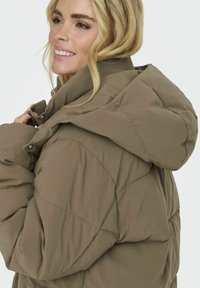 Veste matelassée marron avec capuche, présentant un design matelassé et des poches latérales. Texture douce et lisse avec une coupe décontractée.