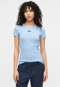 Ανοιχτό μπλε ριμπ μπλούζα με στρογγυλή λαιμόκοψη, κοντά μανίκια και μικρό μπλε λογότυπο "HUGO" στο στήθος. Φοριέται με σκούρο παντελόνι.