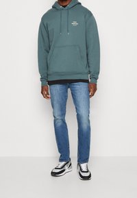 Huvtröja i teal med kenguruficka och vit logotyp, kombinerad med ljusblå jeans och svartvita sneakers.