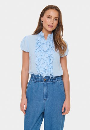 Bluză de culoare albastru deschis cu front volanțat și mâneci scurte tip balon, asociată cu pantaloni din denim cu talie înaltă, având un fermoar și un elastic în zona taliei.