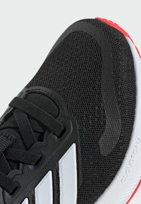 Baskets de sport noires avec une tige en maille, design à trois bandes blanches et semelle rouge. Présente des lacets et des accents texturés pour une adhérence accrue.