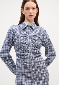 Camicetta a quadretti nei colori blu, bianco e bordeaux; presenta un colletto, maniche lunghe e dettagli arricciati lungo il davanti.