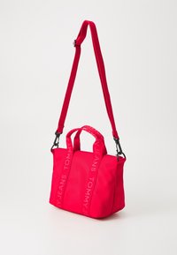Czerwona torba nylonowa z podwójnymi uchwytami i odpinanym paskiem na ramię. Na pasku znajduje się logo "TOMMY JEANS". Kompaktowy i stonowany design.