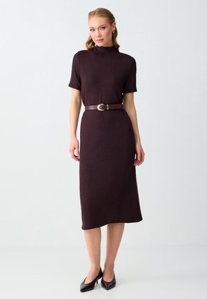 Femme portant une robe midi manches courtes couleur violet foncé avec un col montant et une ceinture marron, associée à des escarpins noirs pointus.