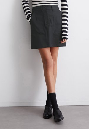 Jupe gris foncé à taille haute, avec deux poches avant et une fermeture à boutons. Portée avec une chemise à rayures noires et blanches à manches longues, des chaussettes noires et des mocassins.