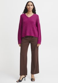 Pull en tricot magenta avec un col en V et des manches longues, associé à un pantalon large marron. Les chaussures sont des sandales à talons hauts noires.