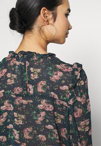 Blusa nera a fiori con motivi rosa e verdi, scollatura arricciata e dettaglio con zip sul retro. Tessuto leggero con una consistenza morbida.