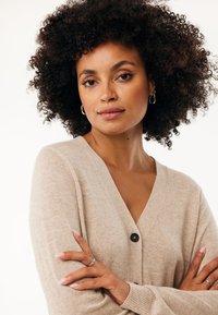 Béžový kardigan s hlubokým V výstřihem, žebrovanými manžetami a jedním černým knoflíkem. Měkká textura s mírným leskem a volný střih.