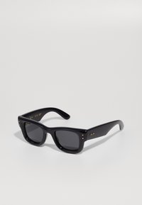 Black sunglasses with a glossy finish, rectangular frame, dark lenses, and gold logo accents on the temples. Two gold dots on each front corner.