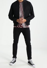 Czarna bomberka nałożona na bordową koszulę w wzory, w zestawie z czarnymi dżinsami i czarno-białymi trampkami. Prosty design bez widocznych logo.