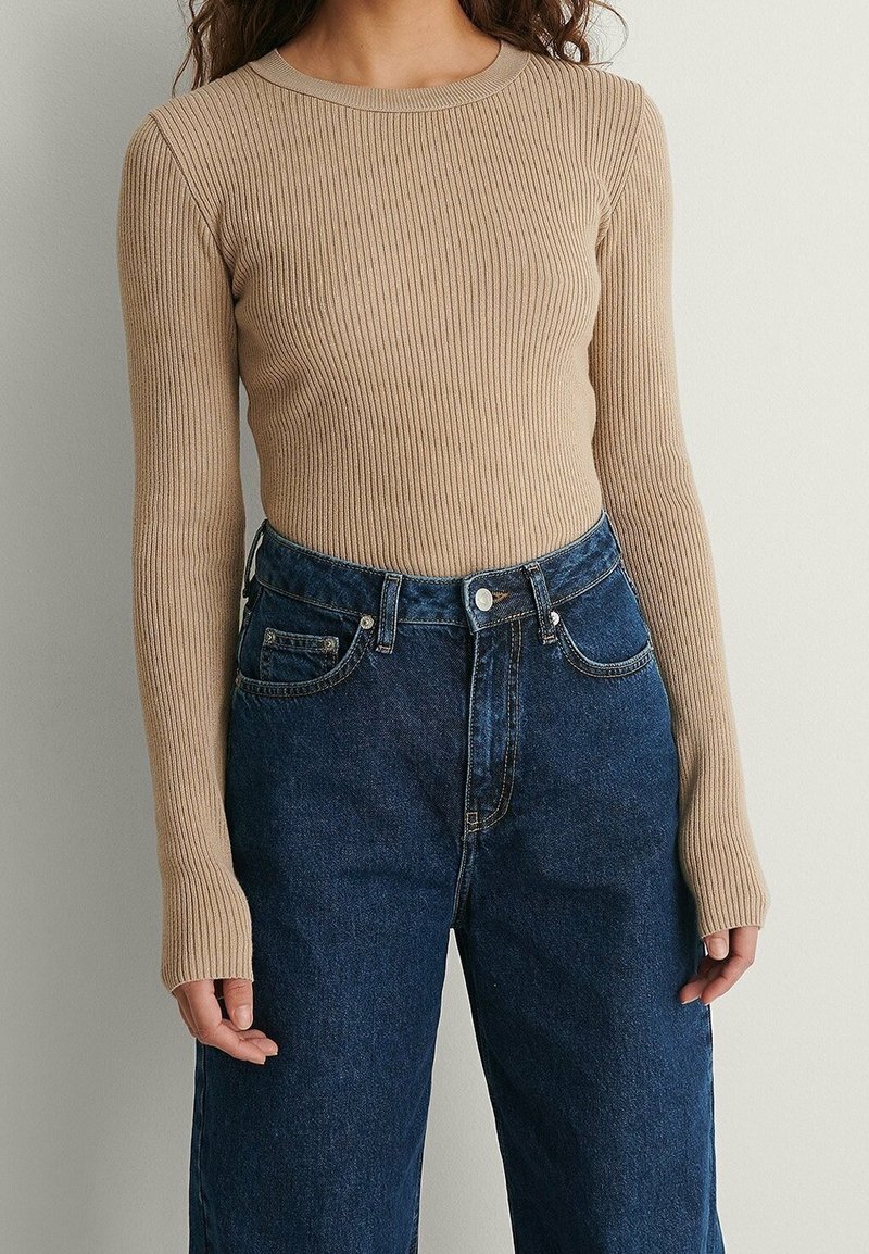 Femme portant un haut beige à manches longues côtelé, rentré dans un jean en denim bleu foncé taille haute, sur un fond clair uni.