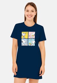 Giovane donna che indossa un vestito blu navy con illustrazioni di Snoopy e Woodstock contrassegnate con le parole felice, amore, affamato e assonnato in scatole colorate.