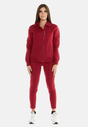 Femme debout portant une veste rouge zippée et un pantalon assorti avec des baskets blanches sur un fond clair uni.