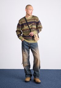 Striktrøje med grønne, brune og blå mønstre; slidte blå jeans med hul; brune støvler; og en lille beige crossbody taske.