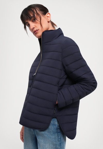 Quilted navy jacket with a high collar, front zipper, and side pockets; features a curved hem and textured finish. Worn over light blue jeans.