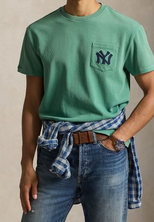 Personne portant un t-shirt vert avec un logo bleu foncé sur la poche poitrine, une chemise à carreaux bleue nouée à la taille, un jean en denim, une ceinture marron et une montre-bracelet.