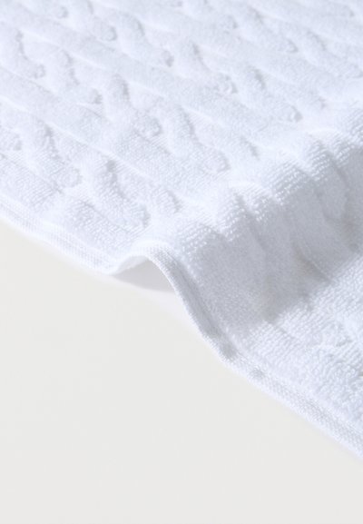 Λευκό υφασμάτινο ύφασμα με ανάγλυφο σχέδιο και διπλωμένη άκρη πάνω σε φωτεινή επιφάνεια, που αναδεικνύει την απαλότητα και τις υφαντές λεπτομέρειες.