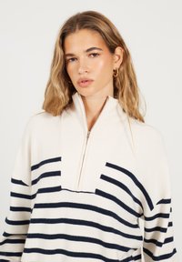 Kobieta z falującymi blond włosami, nosząca złote kolczyki w kształcie kół oraz biały sweter w niebieskie poziome paski z zapięciem na suwak z przodu.