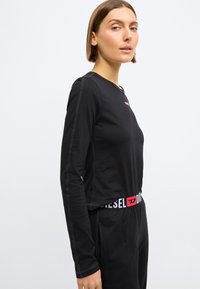 Camisa preta de manga longa, feita de tecido macio, com decote redondo, costuras em contraste e um logótipo "Diesel" vermelho e branco no peito e na cintura.