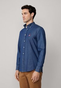Donkerblauwe denim blouse met knopen, voorzien van een kraag, lange mouwen en geborduurd logo. Gladde textuur, comfortabele pasvorm, afgeronde zoom.