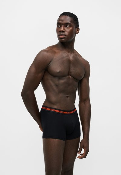 Homme musclé portant un boxer noir avec une ceinture rouge, debout devant un fond clair uni, regardant sur le côté.