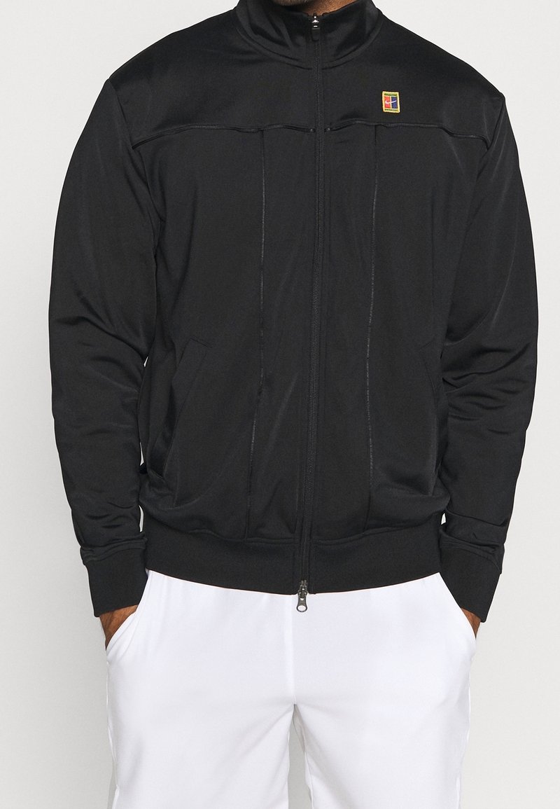 Homme portant une veste noire zippée avec un petit logo coloré sur la poitrine et un pantalon blanc, mains dans les poches, vue de face recadrée au niveau du cou.