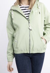 Kvinna iklädd en ljusgrön zip-up jacka med huva och vita snören, randig tröja under, samt mörkblå jeans, med händerna i fickorna.