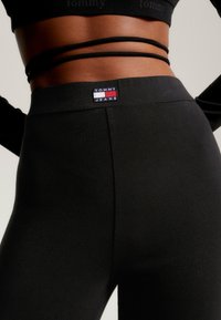 Černé legíny s vysokým pasem, na kterém se nachází obdélníková cedulka "TOMMY JEANS". Materiál vypadá hladce, má přiléhavý design a minimální švy.