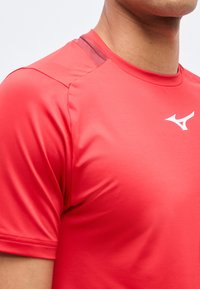 Εγγύς λήψη ενός ατόμου που φοράει κόκκινο αθλητικό πουκάμισο με μικρό λευκό λογότυπο στο στήθος, εστιάζοντας στις λεπτομέρειες του ώμου και του λαιμού.