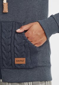 Hoodie bleu marine avec poche en tricot à motif en relief et cordon de serrage ajustable. Comprend une étiquette en cuir avec le branding "oxmo". Texture douce.