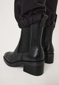 Botas de tobillo de cuero negro con un acabado texturizado, paneles laterales elásticos y un tacón cuadrado. El diseño presenta acentos de hardware mínimos.