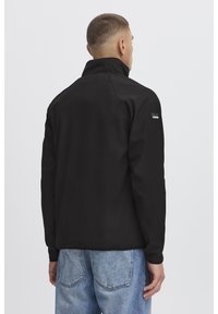 Zwarte jas met hoge kraag, gladde stof en een klein logopatch op de linker schouder. Gepaard met lichtblauwe spijkerbroek.