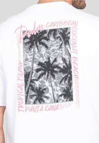 T-shirt blanc avec un imprimé graphique de palmiers au-dessus de l'eau. Le texte comprend "Palm Caribbean" et "Punta Cana" en rose et noir.