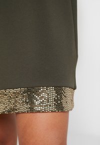 Oliwkowa sukienka wykończona dekoracyjną taśmą ze złotych i srebrnych cekinów, z gładkiego, teksturowanego materiału i prostym krojem.