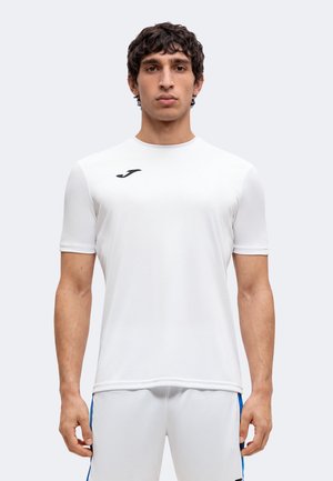 Homme portant un t-shirt de sport blanc uni à manches courtes avec un logo noir et un short blanc à rayures bleues, debout devant un fond blanc.