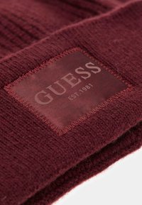 Gorro de punto burdeos con un patrón texturizado y un parche de cuero rectangular que presenta "GUESS EST 1981" en letras en relieve.