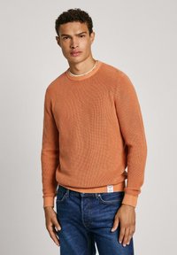 Oranžna rebrasta pulover s okroglim izrezom, z dolgimi rokavi in teksturiranim zaključkom, v kombinaciji s temno modrimi kavbojkami.