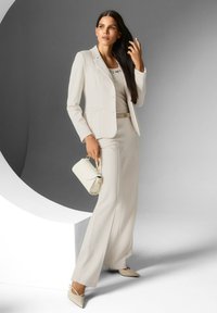 Vrouw met lang donker haar, gekleed in een crèmekleurige blazer, bijpassende wijde broek, beige hakken en een kleine crèmekleurige handtas, poseert binnen.